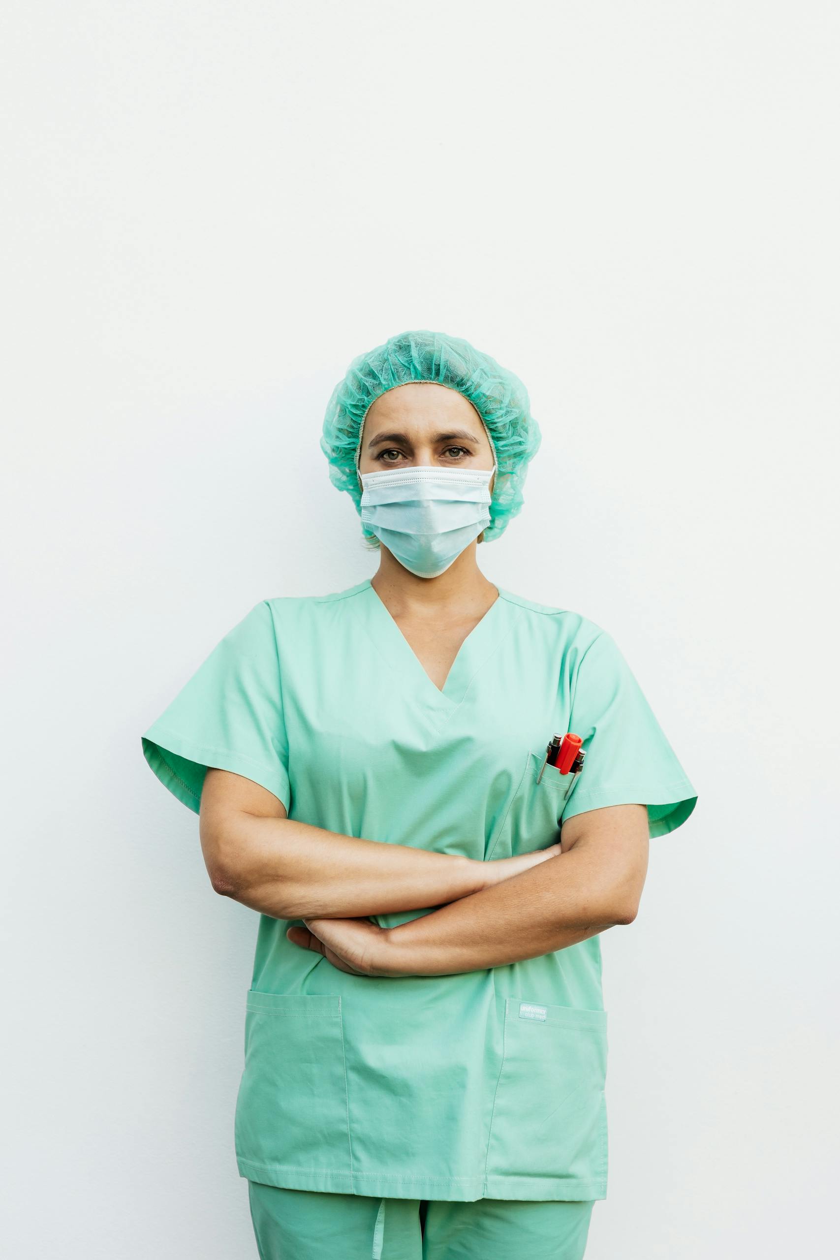 A healthcare professional standing with arms crossed in green scrubs and a protective facemask. How to Become a Travel Nurse